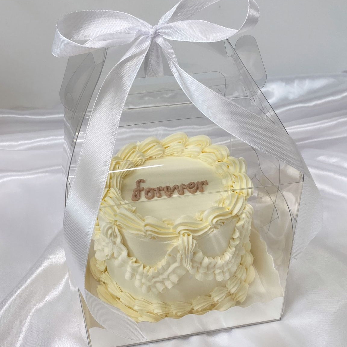 Small decorative cake with 'forever' text in a clear box with a white ribbon on a white background